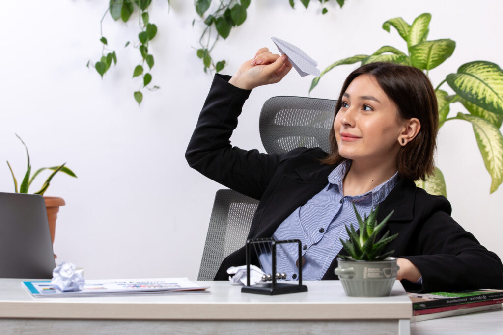 front-view-beautiful-young-businesswoman-black-jacket-blue-shirt-smiling-throwing-paper-planes-front-table-business-job-office-scaled