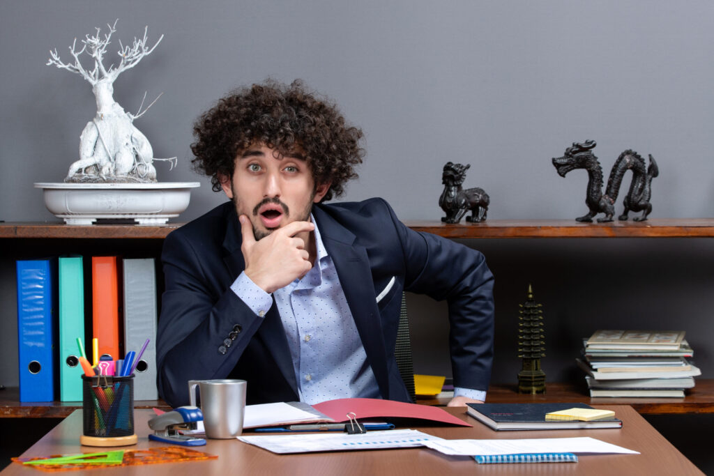 front-view-wondered-businessman-putting-hand-his-chin-sitting-desk-office-scaled